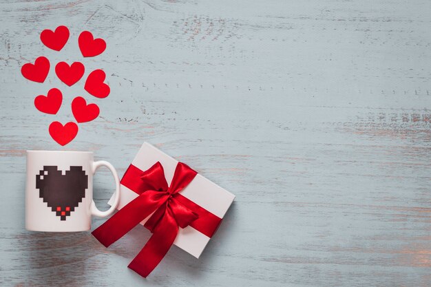 Paper hearts, mug and a white present with red ribbon on a light painted wooden background. Top side angle view, flat lay. Valentines day concept. Copyspace.