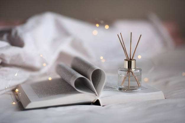 Paper open book with floded pages in heart shape in bed with bottle of liquid home scented perfume and bamboo sticks over glow lights Cozy hygge atmosphere Aromatherapy