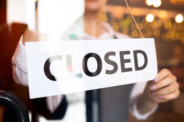 Paper with notice closed being hung by young waitress of classy restaurant or cafe standing behind the door at the end of working day