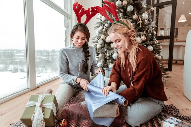 Paper for wrapping. curious hard-working ladies sitting on\
carpet in living room and covering boxes for friends and relatives\
christmas concept