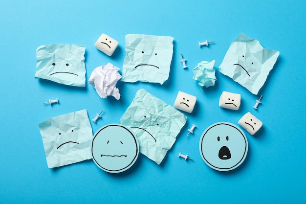 Papers and marshmallows with sad emoji on blue background top view