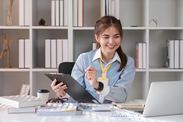 Photo paperwork happy smiling asian business woman in formal wear sitting at wooden desk in modern office and reading report document