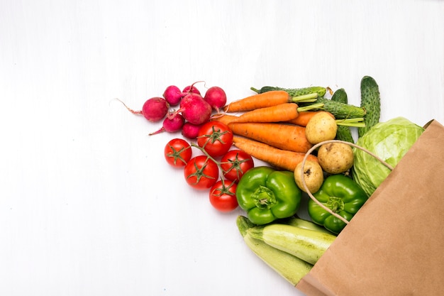 Papieren boodschappentas en verse biologische groenten op een witte houten oppervlak. concept van het kopen van boerderij groenten, het verzorgen van gezondheid, vegetarisme. landelijke stijl, farm fair. plat lag, bovenaanzicht