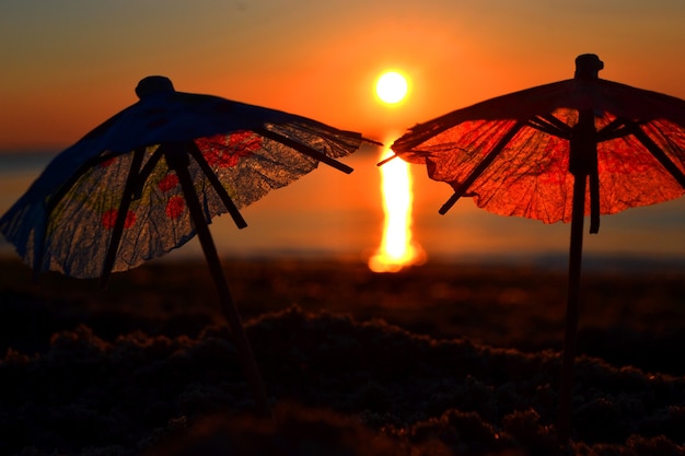 Papieren cocktailparaplu's in zand aan de kust bij zonsondergang, close-up