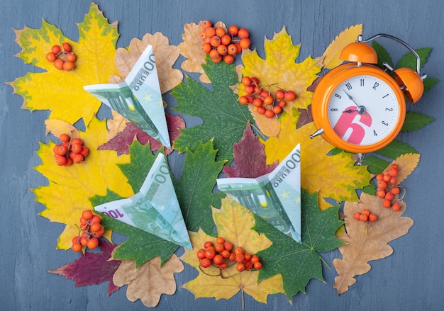 Papieren vliegtuigen gemaakt van eurobankbiljetten en een wekker op een blauwe achtergrond met rode en gele bladeren en oranje bessen. Herfst tijd voor reizen. Reisconcept