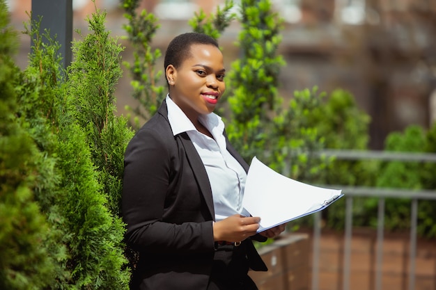 Papierwerk. Afro-Amerikaanse zakenvrouw in kantoorkleding glimlachend, ziet er zelfverzekerd en gelukkig, druk uit. Financiën, zaken, gelijkheid en mensenrechtenconcept. Mooi jong vrouwelijk model, succesvol.