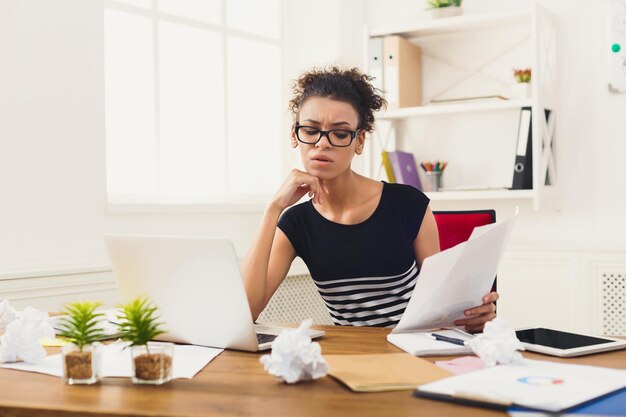 Papierwerk. Ernstige zakenvrouw in formele kleding die aan een houten bureau in een modern kantoor zit, rapportdocument leest, kopieerruimte