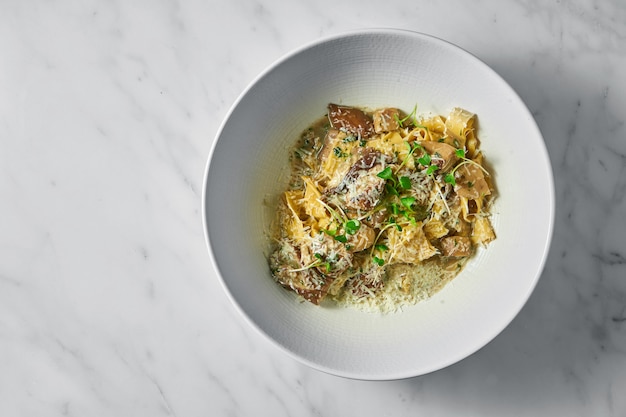 Pappardelle pasta with white forest mushrooms, mushroom sauce, grated parmesan and microgrine in a white bowl on white marble