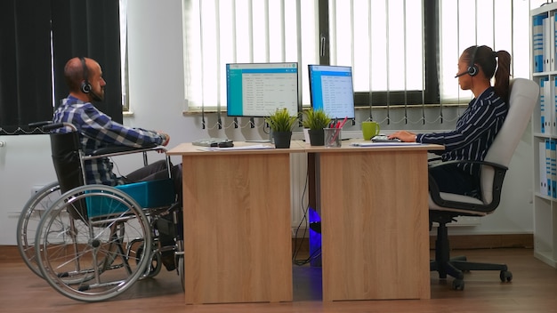 Uomo d'affari paralizzato in sedia a rotelle utilizzando l'auricolare che fa telemarketing e offre assistenza clienti. libero professionista disabile immobilizzato che lavora in un edificio aziendale finanziario utilizzando la tecnologia moderna