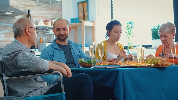 Paralyzed grandfather celebrating his birthday with his family sitting at the kitchen table. Happy couples smiling, drinking wine and eating during a gourmet meal, enjoying relaxing time