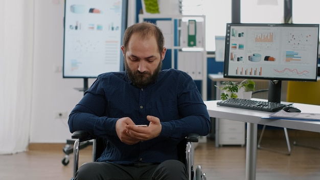 Paralyzed immobilized disabled entrepreneur holding smartphone texting, browsing during work time sitting in wheelchair taking break