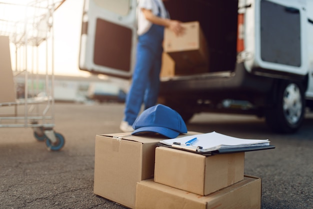 Parcel boxes and cap, deliveryman, delivery service. Man standing at cardboard packages in vehicle, male deliver, courier or shipping job