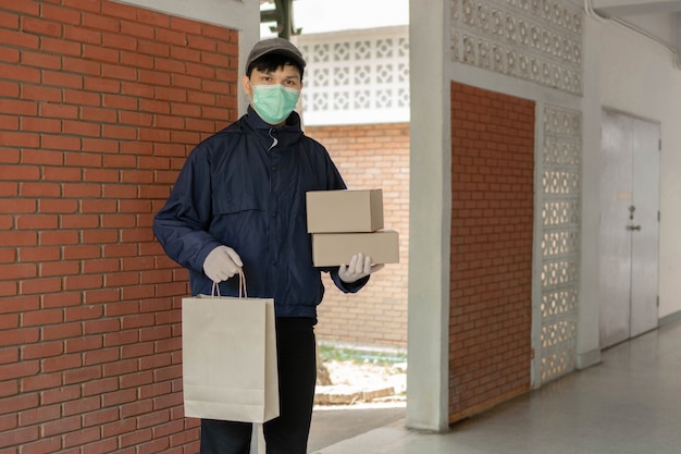 Parcel delivery concept the standing-tall post carrier who is wearing pale grey cap carrying one bag on left hand and two boxes on right hand and being ready to drop these stuffs to his customers.