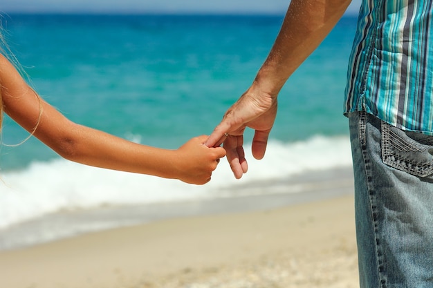 The parent holds the child's hand on the beach in summer