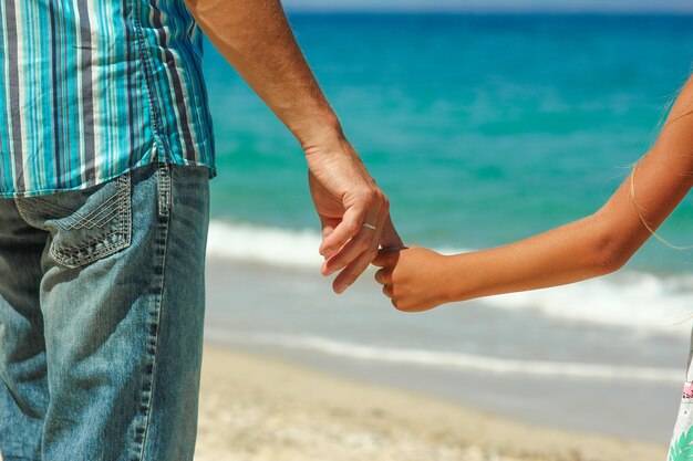 The parent holds the child's hand on the beach in summer