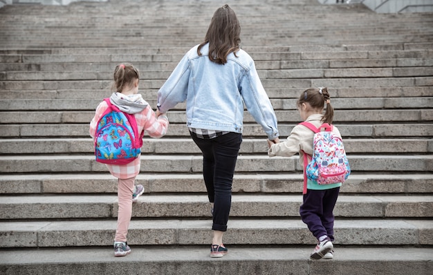 Parent and pupil of primary school go hand in hand. Mom of two girls with a backpack behind the back. Beginning of lessons. First day of fall.