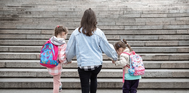 Parent and pupil of primary school. Mom of two girls with a backpack behind the back. Beginning of lessons. First day of fall.