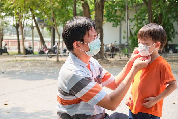 Parent putting mask on his Asian toddler boy child, Dad and son wearing protective medical mask in public area