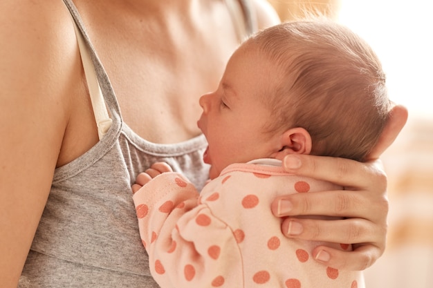 Parenting and new life, faceless mother holding newborn baby in her arms, unknown female wearing gray sleeveless t shirt posing with her tiny kid, child yawning.