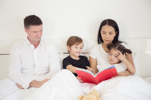 Parents are teaching their daughter and son to read a book in the bedroom on a white bed