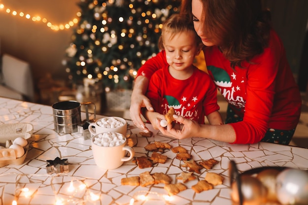 Photo parents and a child in red pajamas prepare christmas cookies in the decorated kitchen of the house