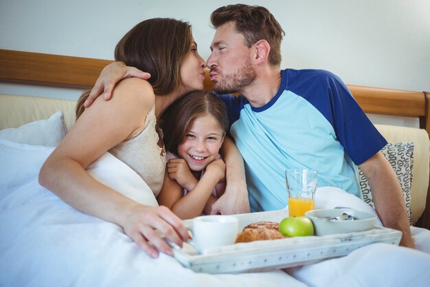 Parents kissing while sitting on bed with daughter and having breakfast