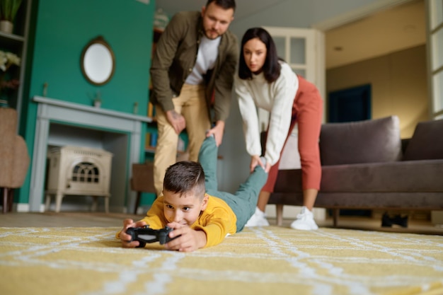 Parents struggle with child addicted to video game. Mother and father holding and pulling boy leg trying to get him out. Internet, digital gadget and technology addiction