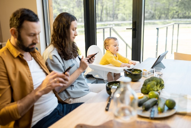 Photo parents and their one year baby boy sitting in mobile devices during lunch at home