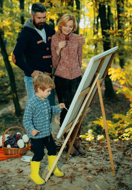 両親は秋の美しい黄金の森公園芸術共同でキャンバスに子供の絵の具を描く息子を見てください。