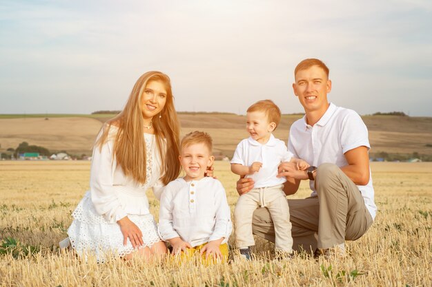 흰 옷을 입은 작은 아들을 둔 부모는 노란 수확된 밀밭에 앉아 푸른 하늘 아래 시골 풍경을 배경으로 덥고 화창한 날 미소를 지으며 휴식을 취합니다.