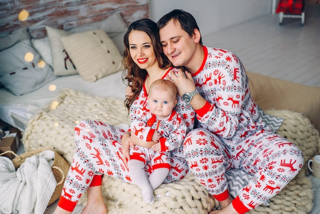 Родители с маленькой дочкой в праздничной одежде с печатными оленями и снежинками сидят на кровати