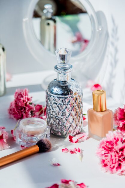 Parfum, foundationcrème, penseel en reflectie met dianthusbloemen op witte muur. make-up concept