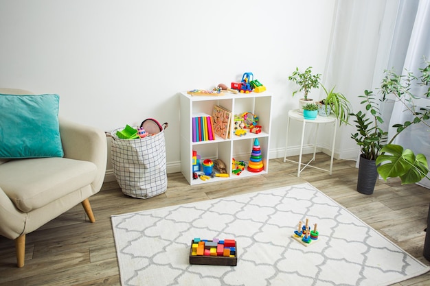 Photo part of children's room with soft beige armchair white shelf for books and toys wooden floor with light rug few toys on a floor