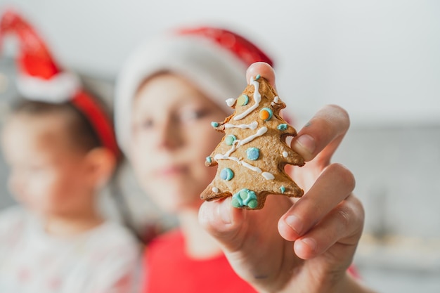 赤いサンタの帽子をかぶった少年の部分的にぼやけた手は、彼によって飾られたクリスマスツリーの形をしたジンジャーブレッドクッキーを示しています