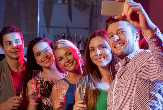 Partij, vakantie, technologie, nachtleven en mensenconcept - glimlachende vrienden met glazen champagne en smartphone die selfie in club nemen