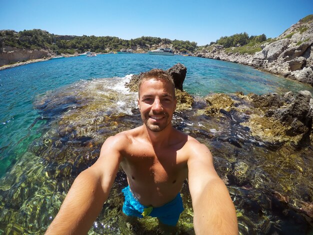 Partly blurred image because of drops of water. Young male explorer in adventure ready for snorkeling. Selfie shot at summer day at rock in the middle of the sea.