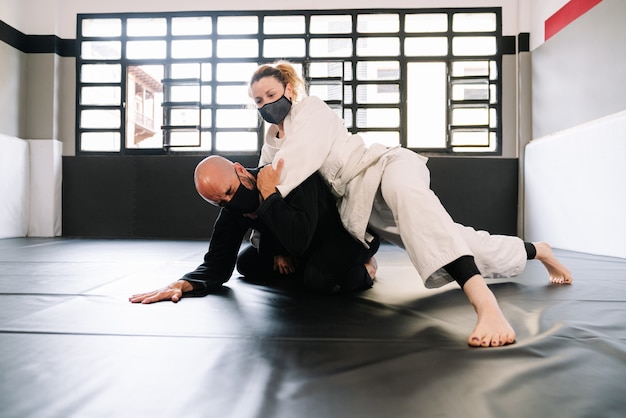 партнеры по боевым искусствам в кимоно, практикующие техники на спортивном коврике, все в масках из-за covid 19