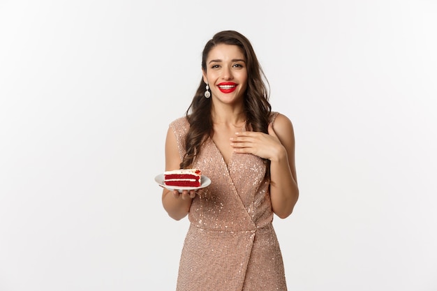 Party and celebration concept. Attractive slim woman in elegant dress holding piece of cake and smiling, standing over white background