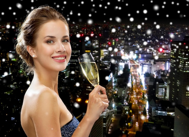 party, drinks, holidays, luxury and celebration concept - smiling woman in evening dress with glass of sparkling wine over snowy night city background