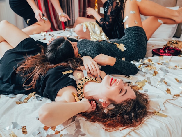 Party girls. Fun and joy. Glitter confetti. Young women in black relaxing on bed. Festive mood and decor. Toothy smiles.