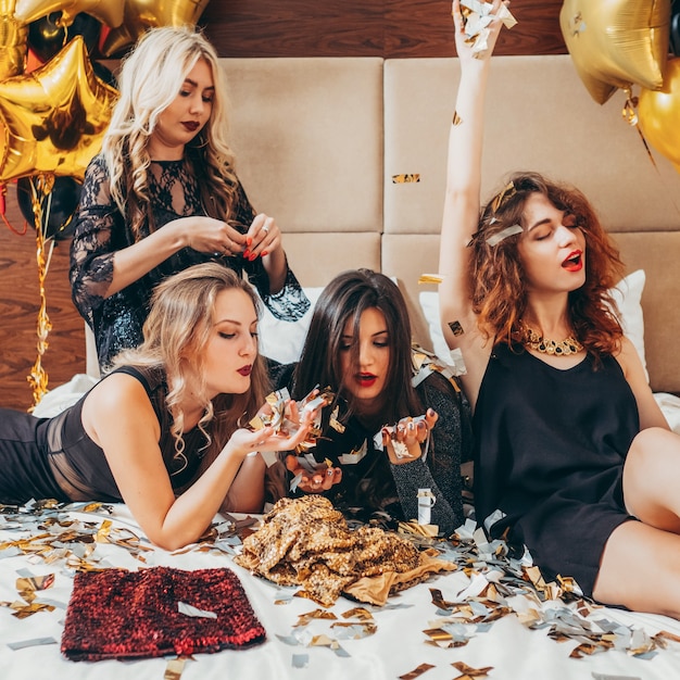 Party girls. Fun and joy. Glitter confetti. Young women in black relaxing on bed. Festive mood and decor