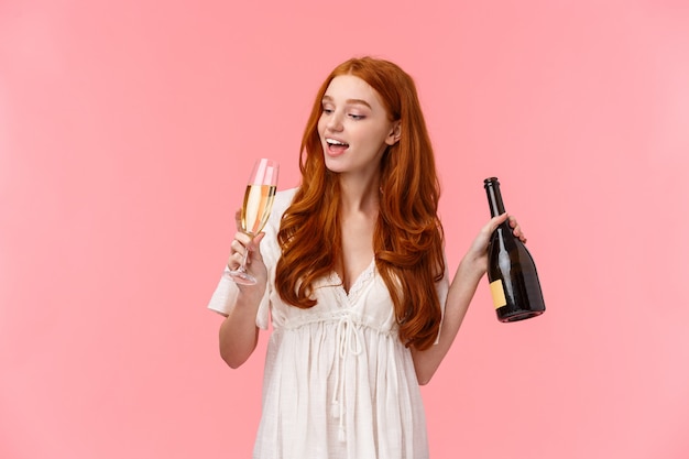 Party, hungover and celebration concept. Happy and cheerful, carefree good-looking redhead woman drinking champagne from glass, holding bottle, got drunk, wasted standing over pink 