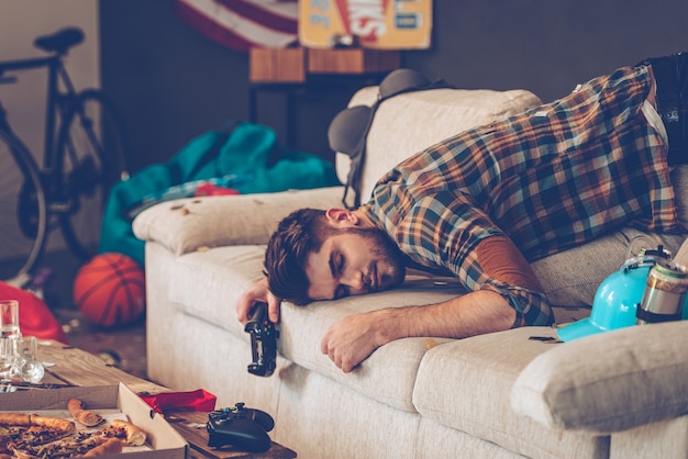 Party is over. Young handsome man passed out on sofa with joystick in his hand in messy room 