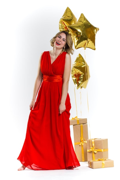 Party photo of elegance sexy lady in red dress with red lips and blond beautiful curly hair, smiling. Background of gold air balloons and gifts. Concept of the sale