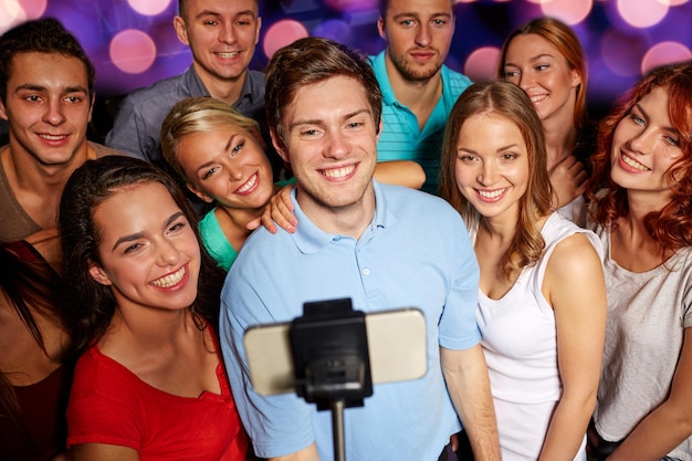 party, technology, nightlife and people concept - smiling friends with smartphone and monopod taking selfie in club