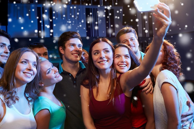 вечеринка, технологии, ночная жизнь и концепция людей - улыбающиеся друзья со смартфоном, делающие селфи в клубе и эффект снега