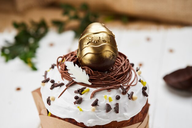 Pasen-cake door gouden chocolade-ei wordt verfraaid op witte houten achtergrond, Traditionele Kulich, Paska klaar voor viering die