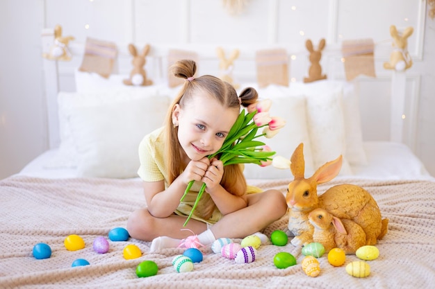 Pasen een klein meisje met geverfde gekleurde eieren en een konijn met lentebloemen tulpen thuis in een lichte kamer die zich voorbereidt op de vakantie die lacht en plezier heeft