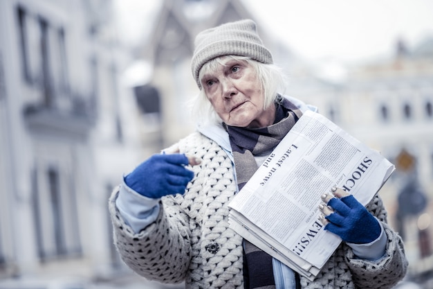 Do not pass by. Sad serious woman selling newspapers while standing on the street