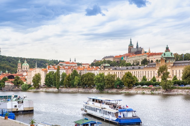 チェコ共和国プラハの歴史的な街並みと曇り空を背景に川沿いを移動する客室クルーザー。